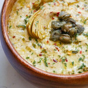 Creamy Artichoke Sauce with Capers in a wooden bowl on a white countertop with tarragon and red chili pepper garnish.