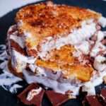 S'mores French Toast cut in half on a black circular stone plate with chocolate and fluff spread
