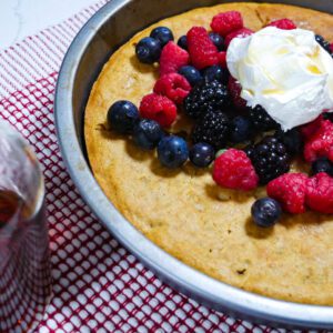 Tiger nut flour oven cake topped with berries and whipped cream in a cake pan, on a red and white placement with maple syrup in a bottle on the side.