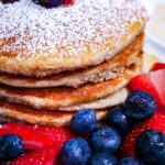 Tiger Nut Flour Pancakes. Blueberries and strawberries around the base and powdered sugar and blueberry sauce on a stack pancakes on a white plate.